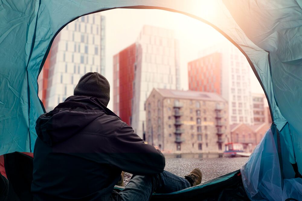 A person wears a jacket and toque and sits in a tent. The tent door is open and looks out to city buildings.