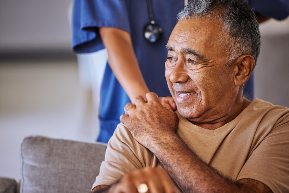 A healthcare worker places a hand on a man's shoulder who sits in a chair. He smiles and has his hand on top of the healthcare worker's hand.